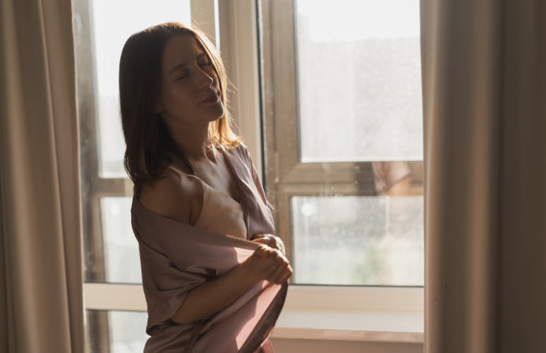 Soft portrait of a woman standing by a window in gentle morning light, reflecting on her body after childbirth