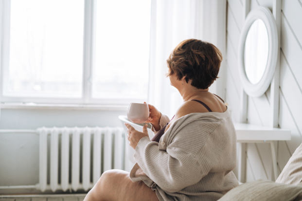 A woman in her late 40s sitting by a window with soft morning light, reflecting calmly — representing the natural transition of menopause and the importance of awareness, care, and balance.