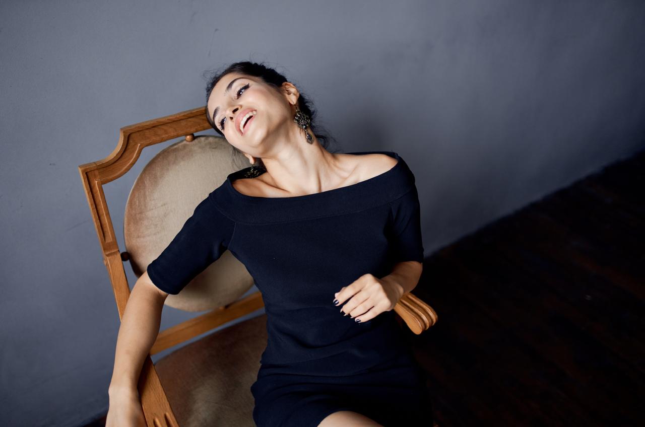 A woman in a black off-shoulder dress leans back in a chair, smiling softly with her head tilted. Her relaxed posture contrasts with an underlying theme of hormonal imbalance, reflecting how mood and energy can fluctuate even when someone appears calm and composed on the outside.